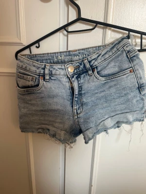 Ljusblå jeansshorts från LAB Industries - Säljer ett par ljusblå jeansshorts från LAB Industries. De har råa, fransiga benslut, klassisk femficksdesign och knappgylf. Perfekta för varma dagar och har en avslappnad, loose passform.