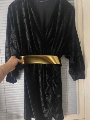 Svart sammetklänning med guldskärp - Snygg svart kort sammetklänning från Nelly med omlott-v-ringning och trekvartsärm. Klänningen har ett brett metallicguldigt bälte i midjan som ger en cool kontrast. Perfekt för dig som vill sticka ut med en unik look.