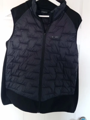 Svart dunväst från Everest - Snygg svart dunväst från Everest med quiltat mönster och dragkedja framtill. Västen har en sportig look med mjuka sidopaneler i stretchmaterial och diskret logga på bröstet. Perfekt för lager-på-lager och outdoor-aktiviteter.