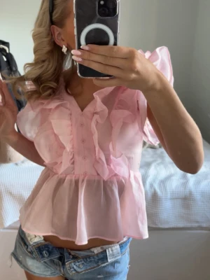 Rosa volangblus med v-ringning - Supersöt rosa blus med v-ringning, volanger längs axlar och framstycke samt knappar framtill. Jag säljer blusen för att det är för liten.Blusen är gjord i ett tunt, transparent material och har korta, luftiga ärmar. Perfekt för dig som gillar romantisk och lekfull stil. Den har en liten liten fläck på sidan som knappast syns. OBS! Skriv om ni vill ha bild på det