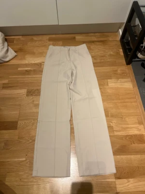 Beige kostymbyxor från Madlady - Snygga beige kostymbyxor från Madlady med raka ben och pressveck. Byxorna har hög midja och stängs med dragkedja och knapp. Perfekta för en clean och stilren look. Materialet känns mjukt och följsamt.