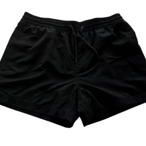 Svarta träningsshorts med snörning - Svarta shorts fran Visual Clothing by MQ med elastisk midja froch snörning framtill. De har en bakficka med knapp och meshfoder på insidan, vilket gör dem perfekta för träning eller bad. Klassisk och enkel design som passar till det mesta.
