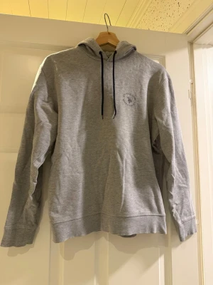 U.S. Polo Assn Grå hoodie - Grå hoodie från U.S. Polo Assn. i enkel och stilren design. Liten logga på bröstet och svarta snören i huvan. Mjuk och skön i materialet – perfekt till vardag eller en avslappnad outfit.  Bra skick. Säljs då den inte kommer till användning längre.