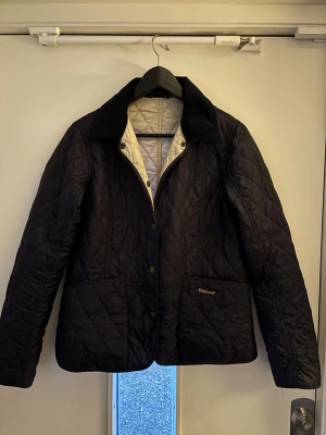Svart quiltad jacka från Barbour - Klassisk svart quiltad jacka från Barbour med krage i manchester och tryckknappar framtill. Jackan har två stora fickor och diskret Barbour-logga på ena fickan. Perfekt för dig som gillar tidlös och stilren design. Pris kan diskuteras 