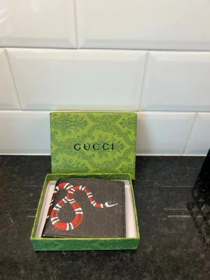 Gucci plånbok med orm-motiv - Snygg plånbok från Gucci i svart canvas med diskret GG-mönster och en rödvit orm som slingrar sig över framsidan. Kommer i original grön Gucci-box. Perfekt accessoar för dig som gillar lyxiga detaljer och unik design.