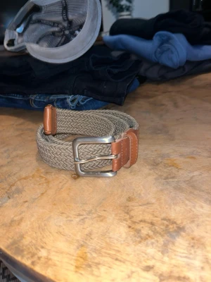 Beige flätat bälte från Jack & Jones - Snyggt beige flätat bälte från Jack & Jones med detaljer i brunt skinn och ett klassiskt metallspänne. Perfekt accessoar för att lyfta din outfit med en stilren touch. Längd 80 cm enligt märkning.