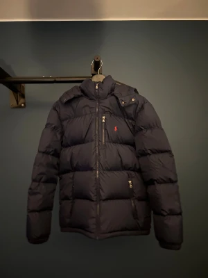 Mörkblå pufferjacka från Polo Ralph Lauren - Säljer en mörkblå pufferjacka från Polo Ralph Lauren med huva och dragkedjor. Jackan har klassisk quiltad design, två sidofickor. Den användes endast ett par gånger förra vintern men är nu för liten. Den är XL barn men sitter som vanlig XS. Priset kan diskuteras! Mvh Alvin 