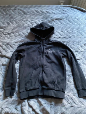 Svart hoodie från Peak Performance för barn  - Säljer en svart hoodie från Peak Performance med dragkedja, två fickor framtill och broderad logga på bröstet. Hoodien har ribbade muddar vid ärmslut och nederkant samt en klassisk huva. Perfekt för chill dagar eller när du vill ha en clean look. Barnstorlek 160
