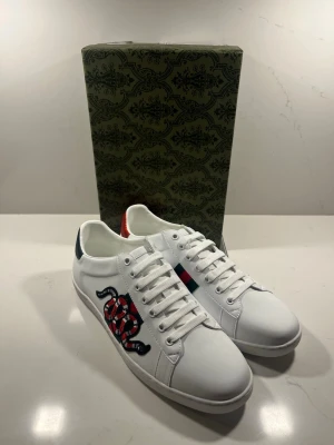 Gucci vita sneakers med orm - Snygga vita sneakers från Gucci med den ikoniska röd-svarta ormen broderad på sidan. Skorna har klassiska gröna och röda ränder på sidan samt metallic-detaljer i rött och grönt på hälen. Tillverkade i slätt läder med vita snören och låg profil.