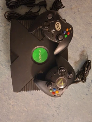Xbox original spelkonsol med handkontroller - Säljer en klassisk svart Xbox original spelkonsol från Microsoft med två matchande svarta handkontroller. Konsolen har den ikoniska gröna Xbox-loggan i mitten och handkontrollerna har färgglada knappar. Perfekt för retrospel och gamingkvällar.
