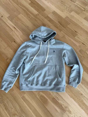 Grå hoodie från Polo Ralph Lauren - Klassisk grå hoodie från Polo Ralph Lauren med broderad marinblå logga på bröstet. Hoodien har vit dragsko, känguruficka och ribbade muddar vid ärmar och nederkant. Tillverkad i mjuk bomullsblandning för maximal komfort.