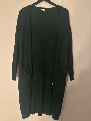 Mörkgrön lång kofta från Vila - Lång, mörkgrön kofta från Vila med två fickor framtill. Koftan har en enkel och stilren design med lång ärm och mjukt stickat material. Perfekt att slänga över en outfit för extra värme och stil.