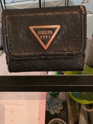 Svart plånbok från Guess - Snygg svart plånbok från Guess med präglat logomönster och en ikonisk trekantig metallogo framtill. Plånboken har en kompakt, fyrkantig form och är tillverkad i konstläder. Perfekt för dig som gillar stilrena accessoarer med tydlig logga. Välanvänd men har mer att ge. 