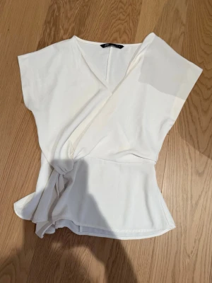 Vit omlottopp från Zara - Säljer en snygg vit omlottopp från Zara med knyt i midjan och v-ringad hals. Toppen har korta ärmar och är tillverkad i ett mjukt material som känns skönt mot huden. Perfekt att styla med jeans eller kjol för en fräsch look.