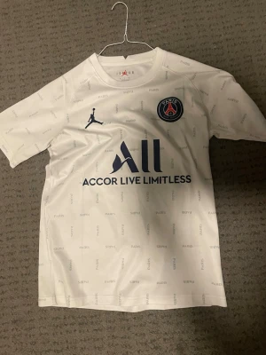 Vit PSG fotbollströja Jordan - Säljer en vit PSG matchtröja i samarbete med Jordan. Tröjan har korta ärmar, diskret Paris-tryck över hela, PSG-logga på bröstet och Jordan-logga. Materialet är lätt och andas, perfekt för träning eller match.
