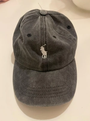 Mörkgrå keps från Polo Ralph Lauren - Snygg mörkgrå keps från Polo Ralph Lauren med den klassiska broderade loggan i vitt framtill. Kepsen har böjd skärm, ventilationshål och justerbar rem bak. Tillverkad i mjuk bomull som ger en avslappnad vibe.