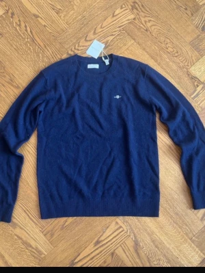 Mörkblå stickad tröja från Gant - Snygg mörkblå stickad tröja från Gant i mjuk lammull. Klassisk rund halsringning och diskret logga broderad på bröstet. Perfekt till jeans eller chinos för en clean och stilren look.