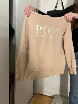 Beige hoodie från Hugo Boss - Snygg beige hoodie från Hugo Boss med stor BOSS-logga i vitt framtill. Klassisk passform med ribbade muddar vid ärmslut och nederkant. Perfekt för en chill och stilren look. Mjuk bomullsblandning som känns skön mot huden.