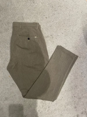 Beige kostymbyxa med rak passform - Snygga beige kostymbyxor med rak passform och klassiska pressveck. Byxorna har bälteshällor, en bakficka med knapp och diskret detalj i metall. Perfekta för en stilren och clean look.