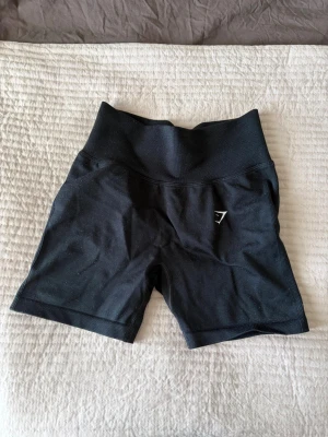 Svarta högmidjade shorts Gymshark - Säljer ett par svarta högmidjade shorts från Gymshark. De är tillverkade i ett stretchigt syntetmaterial och har en tight passform som passar perfekt för träning.