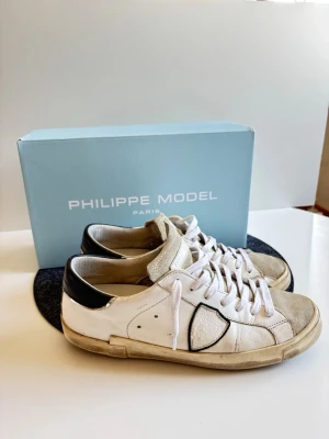 Philippe Model vita sneakers - Hej! Säljer mina snygga vita sneakers från Philippe Model med  och grå mocka på tån. Klassisk låg modell med vita skosnören och diskret logga på sidan. Skorna har en chunky sula och är tillverkade i läder och mocka. Har defekt på hälen (se bild). Philippe modell påse och kartong ingår. Hör av er vid frågor eller funderingar! Pris går att diskutera!