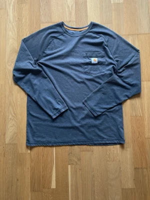 Grå långärmad topp från Carhartt - Snygg mörkblå långärmad topp från Carhartt med vintage look med bröstficka med logga. Tillverkad i mjuk bomull som känns skön mot huden. Perfekt till jeans eller shorts.  Nypris:800kr