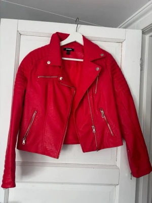 Röd skinnjacka från Junkyard - Snygg röd skinnjacka från Junkyard med coola detaljer som silvriga dragkedjor och tryckknappar. Jackan har en klassisk bikerstil med markerade axlar och quiltade partier på ärmar och rygg. Perfekt för dig som vill sticka ut med en färgstark look.