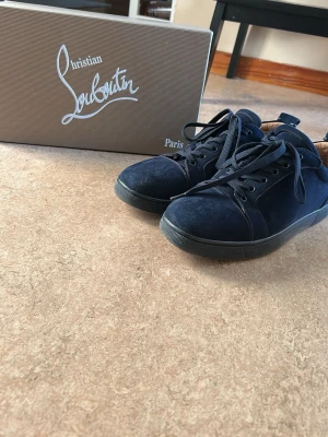 Mörkblå sneakers från Christian Louboutin - Snygga mörkblå sneakers från Christian Louboutin i mocka med klassiska röda sulor och svarta snören. Skorna har en låg profil och stilren design som passar perfekt till streetwear eller en mer dressad look.