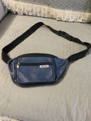 Blå magväska från Wengo - Snygg blå magväska från Wengo i konstläder med svart justerbart band och plastspänne. Väskan har två dragkedjefack fram och ett extra fack på baksidan, perfekt för att hålla koll på småprylar. Enkel och stilren design som passar till det mesta. Helt oskadad!