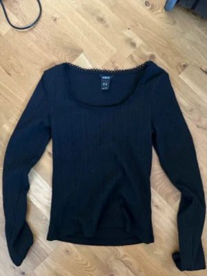 Svart ribbad långärmad topp Lindex - Svart långärmad topp från Lindex med ribbad struktur och rundad halsringning dekorerad med en liten spetskant. Perfekt att styla till jeans eller kjol för en enkel men snygg look.