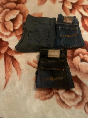Tre par jeans från Nudie och Levi's - Säljer tre par jeans i olika tvättar från Nudie och Levi's. Två par är mörkblå med kontrastsömmar och ett par är klassiskt blått. Alla har bakfickor med signatursömmar och läderpatch i midjan. Perfekta för en avslappnad och trendig look.