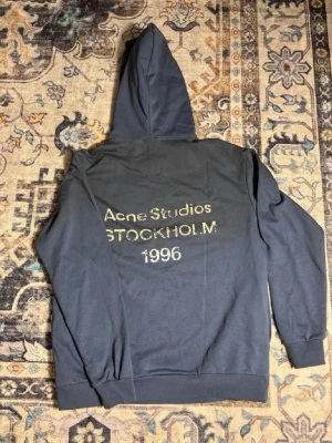 Mörkgrå hoodie Acne Studios - Snygg mörkgrå hoodie från Acne Studios med tryck på ryggen där det står 'Acne Studios STOCKHOLM 1996'. Klassisk huva och ribbade muddar. Perfekt för dig som gillar streetwear och vill ha något stilrent men ändå med attityd.