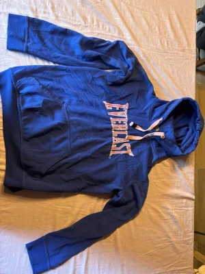 Blå Everlast hoodie med huva - Snygg blå hoodie från Everlast med stor magficka och vit dragsko i huvan. Klassisk logga i vitt och rött framtill. Tillverkad i mjukt bomullsmaterial och har ribbade muddar vid ärmslut och nederkant. Perfekt för en avslappnad och sportig stil.