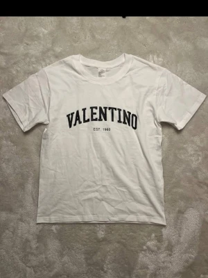 Vit Valentino t-shirt med tryck - Vit t-shirt från Valentino med svart texttryck 'VALENTINO EST. 1960' på bröstet. Klassisk rund halsringning och korta ärmar. Perfekt basic-plagg med stilren design och skön passform.