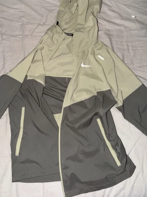 Nike tvåfärgad vindjacka med huva - Snygg vindjacka från Nike i två nyanser av grått och olivgrönt. Jackan har huva, dragkedja framtill och två fickor med dragkedja. Nike-logga på bröstet och reflekterande detalj. Perfekt för en sportig och avslappnad stil.