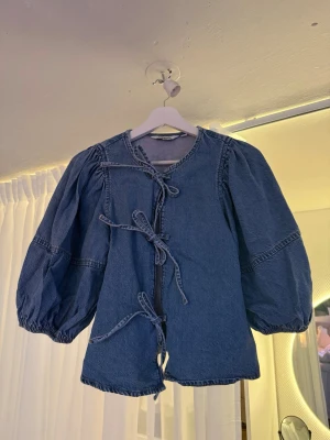 Blå jeansjacka med knytband Kappahl - Trendig blå jeansjacka från Kappahl med unika knytband framtill istället för knappar. Jackan har puffiga ärmar och rund halsringning, vilket ger en cool och modern look. Perfekt för dig som vill sticka ut med en annorlunda jeansjacka.