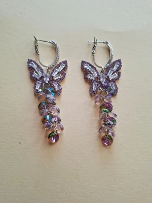 Lila fjärilsörhängen med pärlor - Unika örhängen med lila broderade fjärilar och hängande skimrande pärlor i olika nyanser av lila och transparent. Örhängena har silverfärgade krokar och ger en lekfull och glittrig look. Perfekta för dig som vill sticka ut med detaljerade accessoarer.