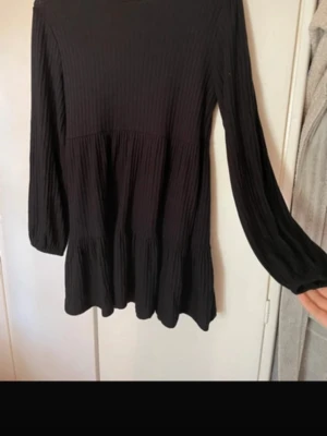 Svart långärmad klänning Chiquelle - Svart långärmad klänning från Chiquelle i ribbad struktur. Klänningen har en lös passform och är tillverkad i ett mjukt material som ger en skön känsla. Perfekt för dig som gillar enkel och stilren design.