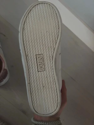 Vita Polo sneakers med glitterdetalj - Snygga vita sneakers från Polo med klassisk låg profil och vita snören. Skorna har en unik detalj med rosa glitter på hälen och insidan, vilket ger dem en extra touch. Tillverkade i läderimitation och passar perfekt till streetwear eller casual outfits.