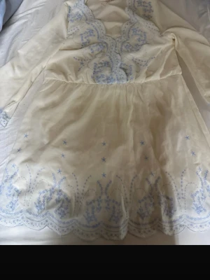 Vit klänning med blå broderier - Söt vit kortklänning med ljusblå broderade blommor och vågig kant nertill. Klänningen har långärmade ärmar och markerad midja. Perfekt för sommarens alla tillfällen och ger en romantisk vibe.