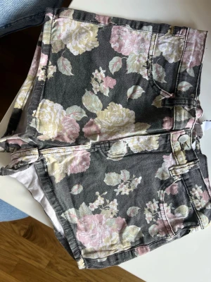 Blommiga shorts med hög midja - Säljer ett par snygga shorts med hög midja och blommigt mönster i pastellfärger som rosa, gul och grön på en mörk bas. Shortsen har klassisk femficksdesign och är perfekta för varma dagar. Passar dig som vill ha en trendig och somrig look.