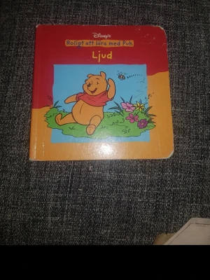 Roligt att lära med Puh: Ljud - Barnbok med Nalle Puh på omslaget, färgglad med röd, orange och blå framsida. Boken heter 'Roligt att lära med Puh: Ljud' och har en illustration av Puh som ligger på gräset bland blommor. Perfekt för dig som gillar klassiska Disney-karaktärer.