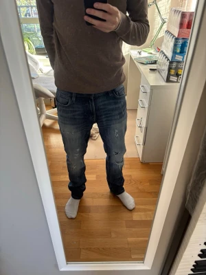 Blå slitna jeans med tvättade detaljer - Säljer ett par blå jeans med slitningar och tvättade detaljer. Jeansen har normal passform, fem fickor och klassisk knappgylf. Perfekta för en avslappnad och trendig look.