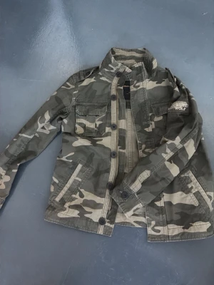 Camouflage vindjacka från Cruel Wolf - Snygg camouflagefärgad vindjacka från Cruel Wolf med klassisk krage, knappar framtill och flera fickor. Jackan har ett coolt militärinspirerat mönster och är tillverkad i bomull. Perfekt för dig som gillar streetwear och utilitystil.