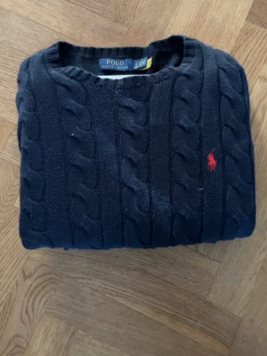 Mörkblå kabelstickad tröja Polo Ralph Lauren - Kabelstickad mörkblå tröja från Polo Ralph Lauren med rund halsringning och den klassiska röda logotypen broderad på bröstet. Tröjan har en tidlös design och är tillverkad i mjukt material som passar perfekt till många olika stilar.
