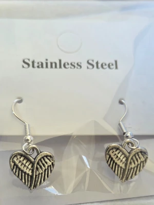 Hjärtfomade örhängen i rostfritt stål - Säljer ett par silvriga örhängen i rostfritt stål med hjärtformade hängen. Hänget har ett detaljerat mönster som påminner om vingar eller fjädrar, vilket ger en unik och cool look. Perfekta för dig som gillar accessoarer med lite edge.