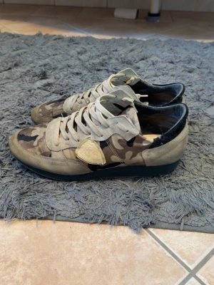 Philippe Model camo  - Snygga sneakers från Philippe Model med camouflagemönster i grönt, beige och brunt. Skorna har beige mockadetaljer, vita skosnören och svart hälparti. Klassisk låg modell med gummisula och ikonisk sköld på sidan. Perfekta för en streetwear-look.