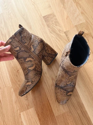 Bruna stövletter med ormmönster - Snygga bruna stövletter med chunky klack och trendigt ormmönster. Dragkedja på sidan för enkel påtagning och cool look. Tillverkade i konstläder med struktur som liknar ormskinn. Perfekta för dig som vill sticka ut med dina skor.