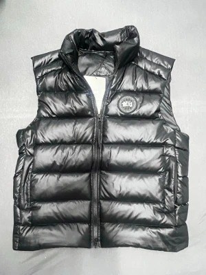 Svart dunväst från Canada Goose - Snygg svart dunväst från Canada Goose med hög krage och dragkedja framtill. Västen har ett quiltat mönster och en rund logotyp på bröstet. Perfekt att slänga över en hoodie eller tröja för extra värme. Klassisk och stilren design som funkar till det mesta. Dessutom finns det NFC 