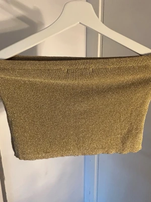 Glittrig guldtubtopp - Snygg axelbandslös tubtopp i guld med glittrigt material. Perfekt för dig som vill sticka ut och addera lite extra bling till din outfit. Stretchigt tyg som sitter tight och bekvämt runt överkroppen.